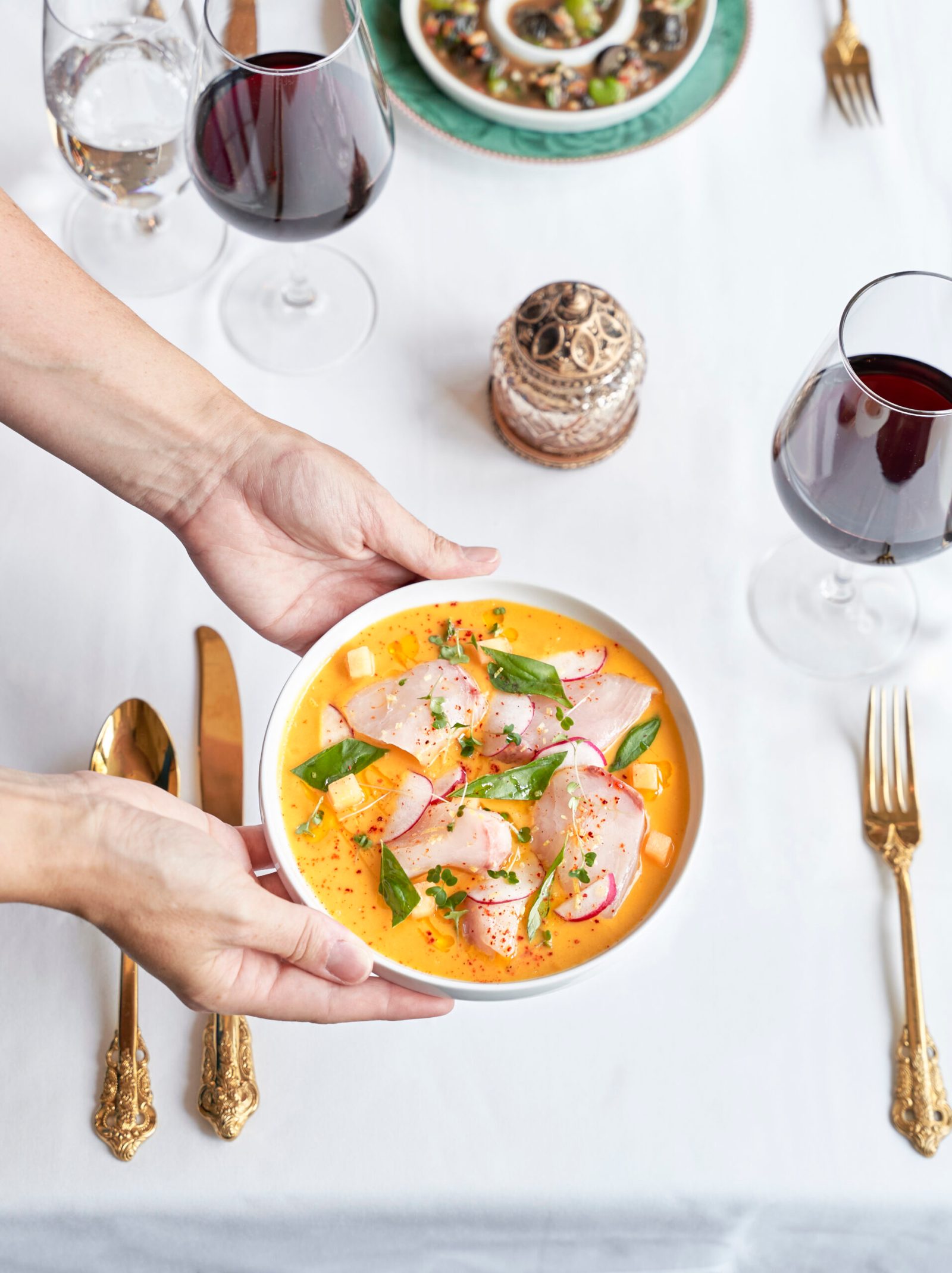 Hands setting down a bowl of cured kanpachi in chilled tomato broth on a white tablecloth