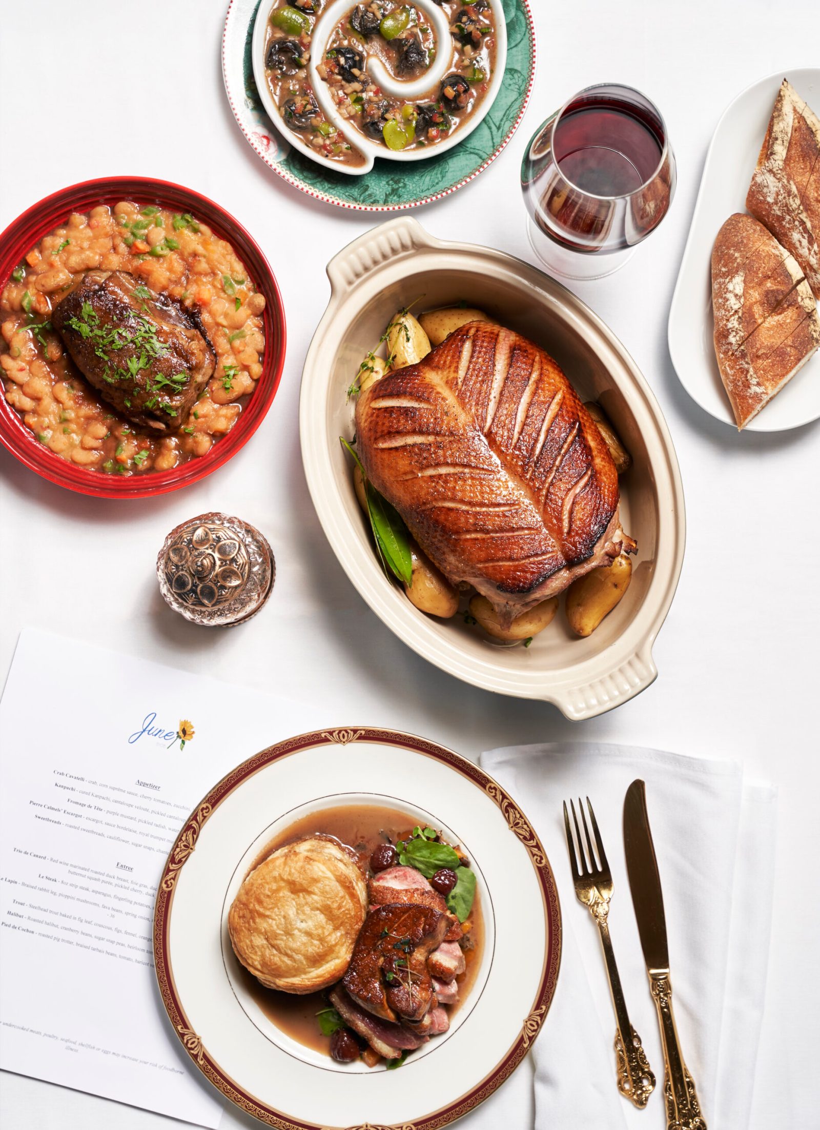 A roasted whole duck in a copper-coloured cocotte, surrounded by escargot, cassoulet, bread and the June BYOB menu